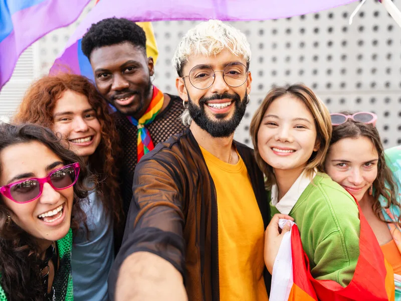 A small group of friends laughing together outdoors, one holding a pride flag low at their side.