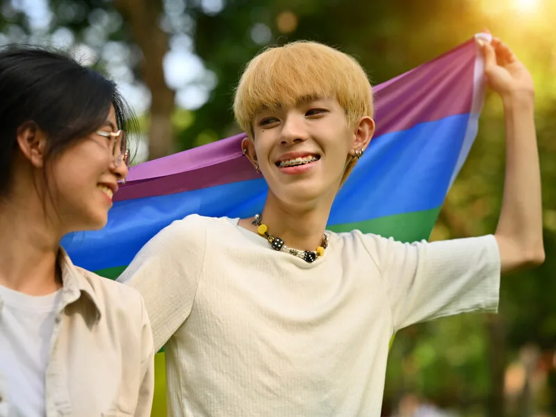 A young person standing close to a friend holding a pride flag, soft afternoon light.