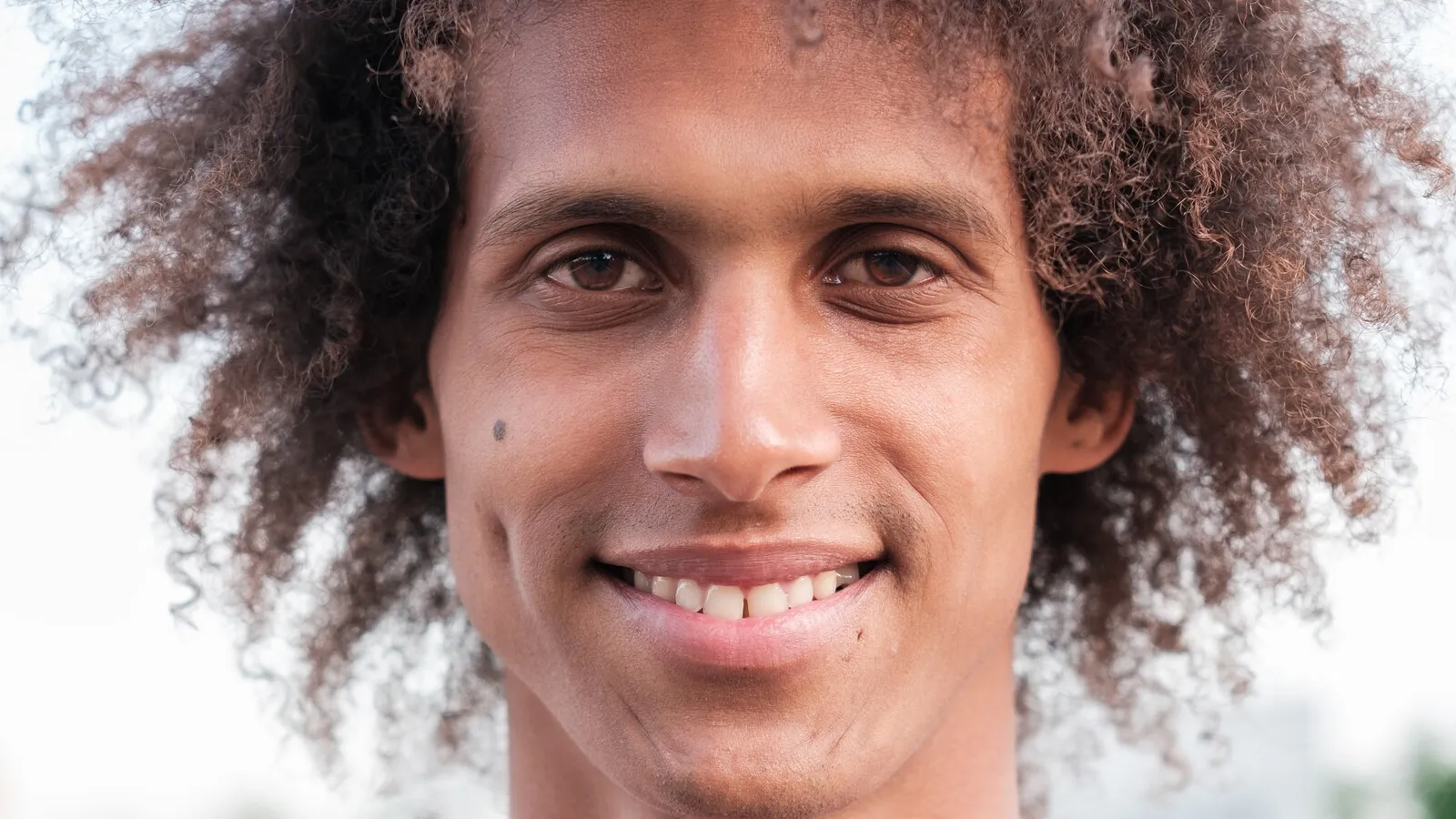 A close-up of a young person with curly hair, smiling softly outdoors in golden afternoon light.