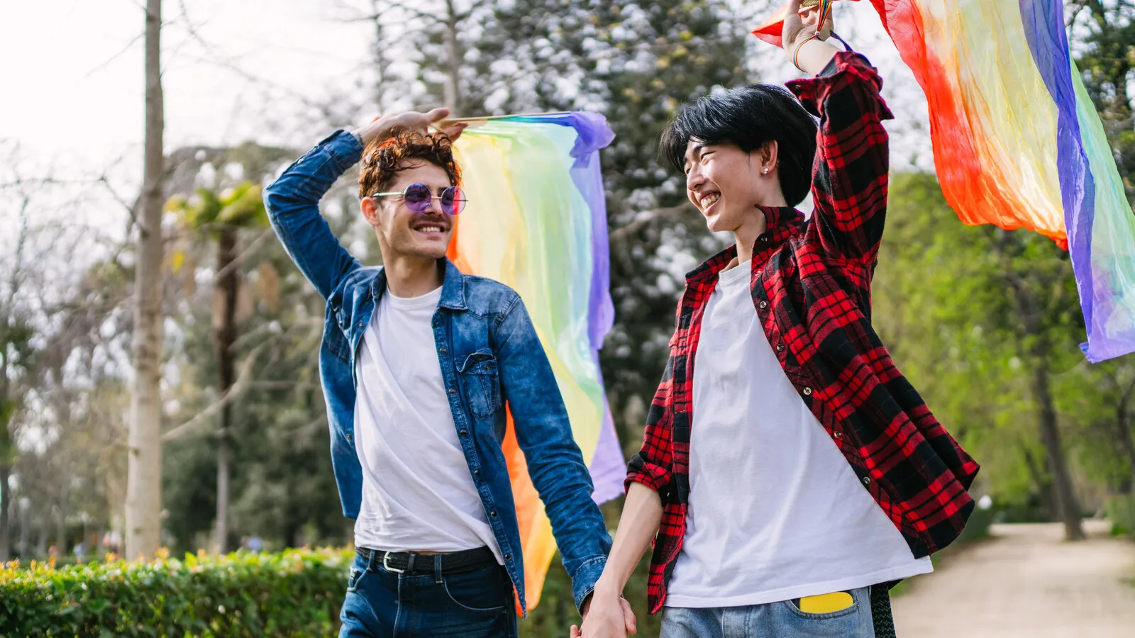A smiling couple holding a pride flag together in a green park, comfortable and warm with each other.