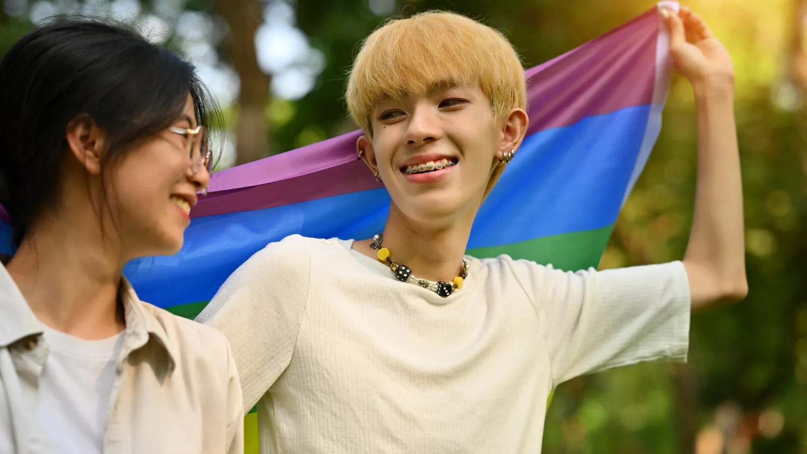 A teenager smiling brightly outdoors while a friend holds a pride flag, both relaxed in afternoon light.